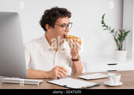 Jeune homme d'affaires avec du tissu mangeant hot dog savoureux dans le bureau Banque D'Images