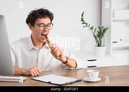 Jeune homme d'affaires mangeant hot dog savoureux au bureau Banque D'Images