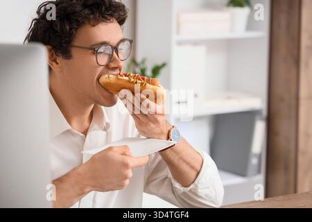 Jeune homme d'affaires mangeant un hot dog savoureux au bureau, gros plan Banque D'Images