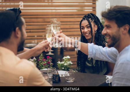 Un groupe diversifié de quatre jeunes amis est assis à une table de restaurant en plein air, parlant, souriant et socialisant par une journée ensoleillée. Banque D'Images