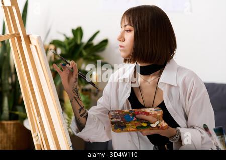 Jeune femme exprime son talent artistique tout en peignant sur un chevalet dans sa maison confortable. Banque D'Images