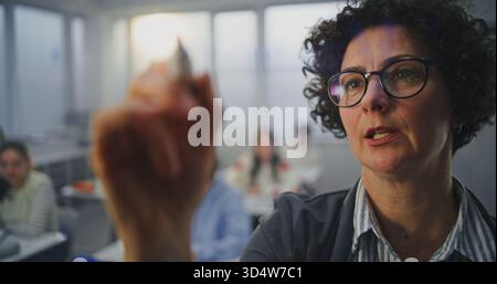 Gros plan femme adulte portant des lunettes écrit activement avec marqueur sur le tableau interactif invisible. Les élèves écoutent attentivement. Concept de méthodes d'enseignement modernes, innovations en technologie éducative. Banque D'Images