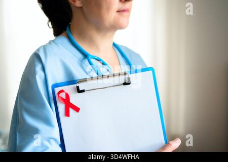 Gentille médecin féminine avec stéthoscope autour du cou et ruban de sensibilisation rouge sur uniforme, souriant chaleureusement pour le soutien et les soins des patients VIH. Banque D'Images