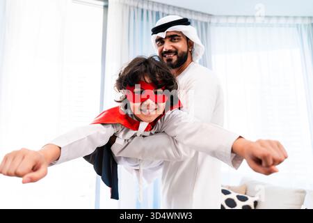 Famille du moyen-Orient portant des vêtements traditionnels émiratis passant du temps ensemble à la maison. Père du moyen-Orient tenant son fils habillé en superher Banque D'Images