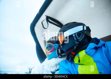 Deux skieurs sont assis sur un télésiège, regardant vers les pistes enneigées. Ils portent du matériel de ski coloré et des lunettes, prêts pour leur prochaine course. Banque D'Images