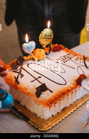 Gâteau d'anniversaire décoré de fruits et de chocolat, orné de bougies allumées et d'ornements floraux. Le gâteau a Buon Compleanno écrit, indiquant un Banque D'Images