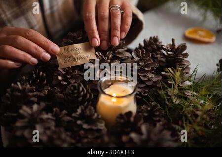 Une scène de vacances chaleureuse montrant des mains arrangeant des pommes de pin autour d'une bougie allumée avec une étiquette manuscrite Joyeux Noël, évoquant la maison, la famille et les fêtes Banque D'Images