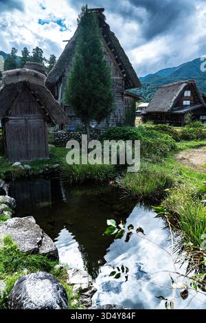Vue des maisons traditionnelles au toit de chaume reflétées dans un ruisseau tranquille sous un ciel spectaculaire, une idylle rurale, Shirakawa, Gifu, Japon. Banque D'Images