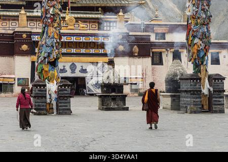 Vue de moines marchant à travers la cour en pierre avec des drapeaux de prière flottant autour des brûleurs d'encens dans l'ancien monastère, Shannan, Tibet, Chine. Banque D'Images
