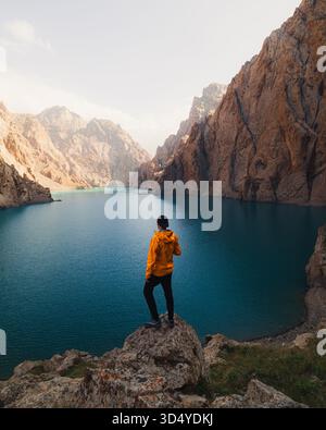 Vue d'une personne dans une veste jaune vif se dresse sur un éperon rocheux surplombant le magnifique lac turquoise entouré de falaises imposantes, Kol-Suu L. Banque D'Images