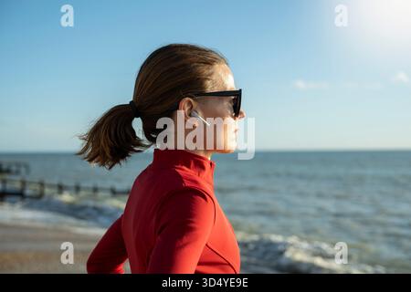 Photo de profil d'une femme sportive portant un vêtement de sport rouge et des lunettes de soleil et des écouteurs sans fil, regardant l'océan Banque D'Images