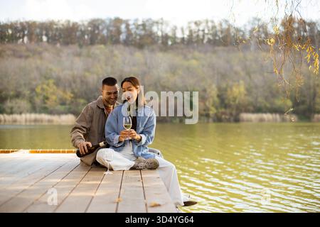 Un couple joyeux est assis sur un quai en bois, partageant un verre et rires tout en étant entouré par la beauté de la nature. Banque D'Images