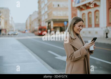 Une jeune femme vêtue d'un manteau beige se tient dans une rue de la ville, souriant en regardant son smartphone. Elle tient une boisson chaude dans son autre main, profitant d'un sunn Banque D'Images