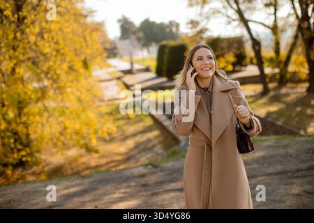Une jeune femme se promène dans un parc pendant la journée, souriant et parlant au téléphone. Les arbres regorgent de couleurs d'automne, créant un bac vibrant Banque D'Images