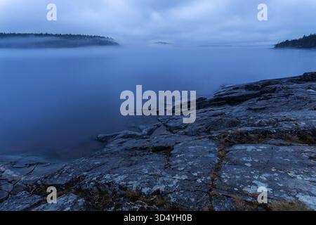 Vue de brouillard roulant sur les eaux tranquilles et sombres, rencontrant le rivage rocheux sous un ciel nuageux, enveloppé de mystère, Sortavala, Russie. Banque D'Images