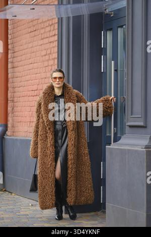 Une femme à la mode dans un long manteau de fourrure brun et des lunettes de soleil sort d'un immeuble de bureaux urbain coloré Banque D'Images