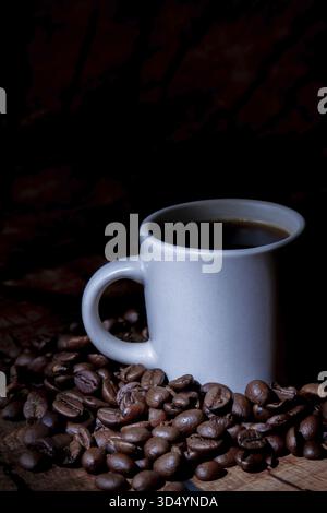 Cuire du café chaud à la vapeur dans une tasse blanche debout parmi des grains d'arabica torréfiés foncés sur une surface en bois brun Banque D'Images