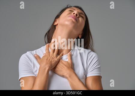 Femme touchant la poitrine et le cou avec les yeux fermés montrant de l'inconfort ou mal de gorge à l'intérieur Banque D'Images