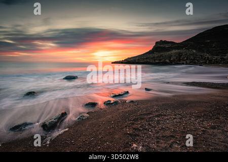 Un lever de soleil époustouflant peint l'horizon tandis que les vagues se répandent sur les rochers et un rivage de galets sombres. Une scène côtière tranquille avec des tons chauds, silhouette de falaise, et Banque D'Images
