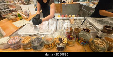 Berlin, Allemagne - 19 septembre 2025 : L'employé d'un magasin de crème glacée fabrique de la crème glacée roulée avec diverses garnitures en vue. Banque D'Images
