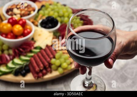 Une main tient un verre de vin rouge tandis qu'un plateau magnifiquement arrangé de viandes, fromages, fruits frais et noix se trouve à proximité. Le réglage est chaud et je Banque D'Images
