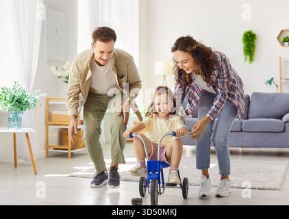 Les parents heureux aident leur fille à monter tricycle à l'intérieur, moment familial ludique à la maison Banque D'Images