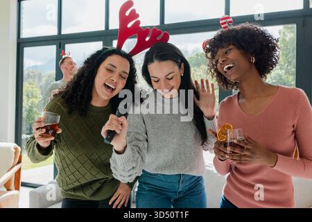 Amis divers chantant karaoké portant des bandeaux en bois tenant un micro et des boissons dans le salon Banque D'Images