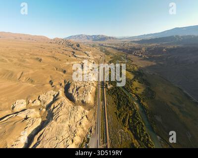 Une vue aérienne à haute altitude d'une autoroute droite et d'une rivière sinueuse coupant à travers une vallée de montagne sèche et aride avec des jaunes texturés fortement érodés Banque D'Images