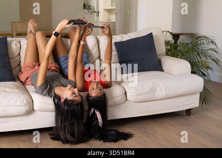 Couché diverses amies faisant défiler des appareils portables sur un canapé blanc cassé à la maison, avec une paume en pot Banque D'Images