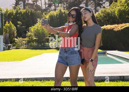 Selfie diverses amies féminines debout près de la terrasse de la piscine arrière, avec smartphone et écouteurs Banque D'Images