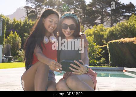 Penchant ensemble diverses amies féminines sur la terrasse en bois à la piscine ensoleillée de la cour arrière, vérifier smartphone Banque D'Images