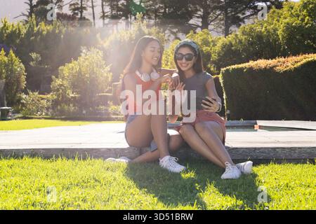 Pointant divers amis féminins assis sur la pelouse dans le jardin à côté de la terrasse, avec des écouteurs de smartphone Banque D'Images