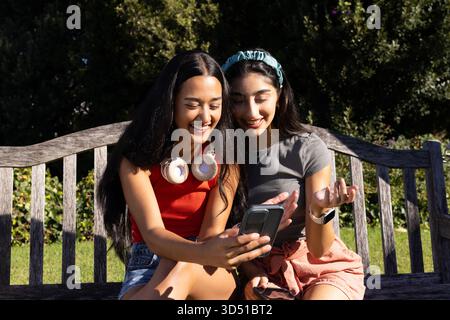 Sourire diverses amies féminines défilant smartphone sur un banc de parc en bois, avec des écouteurs blancs Banque D'Images