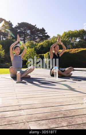 Pratiquer le yoga diverses amies féminines assises sur une terrasse en bois dans le parc, avec des bouteilles et un smartphone Banque D'Images