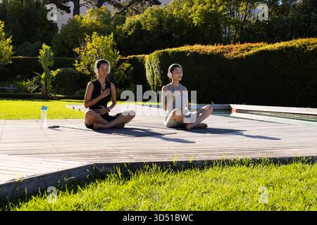 Méditer diverses amies féminines assises les jambes croisées sur le pont près de la piscine, avec des écouteurs Banque D'Images