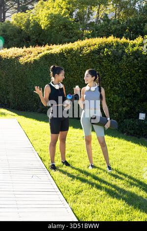 Bavarder diverses amies sur la promenade dans le jardin ensoleillé, tenant des tapis de yoga et des bouteilles d'eau Banque D'Images