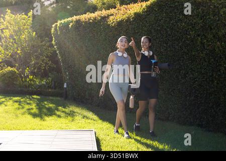 Marcher diverses amies féminines dans la cour avec des tapis de yoga bouteilles d'eau écouteurs sans fil, espace de copie Banque D'Images