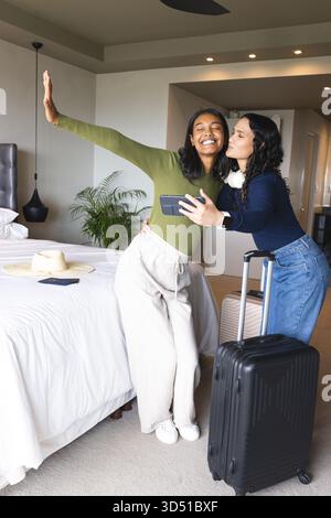 Prise selfie couple féminin diversifié debout dans une chambre d'hôtel moderne, avec smartphone et valise Banque D'Images