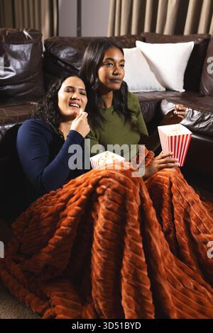 Blotti diverses amies féminines sous une couverture de rouille dans un salon confortable, tenant des seaux de pop-corn Banque D'Images