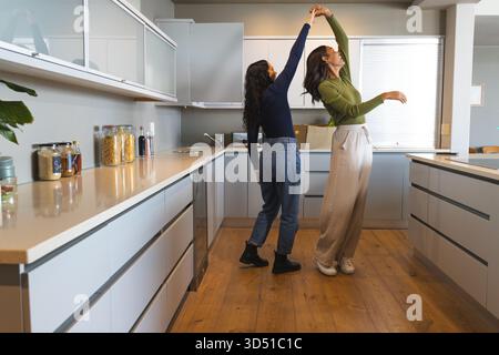 Danser diverses amies féminines se tenant par la main tout en tourbillonnant dans la cuisine moderne, avec des pots en verre Banque D'Images