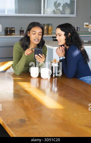 Pointant divers amis féminins regardant smartphone à la table de cuisine, avec tasses et presse française Banque D'Images