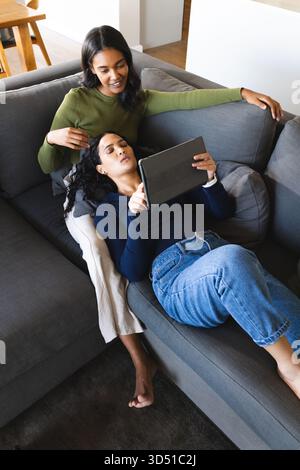 Relaxant diverses amies féminines se prélassant sur le canapé sectionnel gris sur tapis sombre à la maison, avec tablette Banque D'Images
