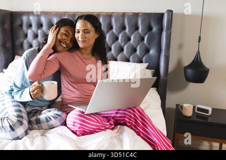 Assis divers partenaires féminins tenant des tasses et utilisant un ordinateur portable sur le lit dans la chambre, avec une lumière pendante Banque D'Images