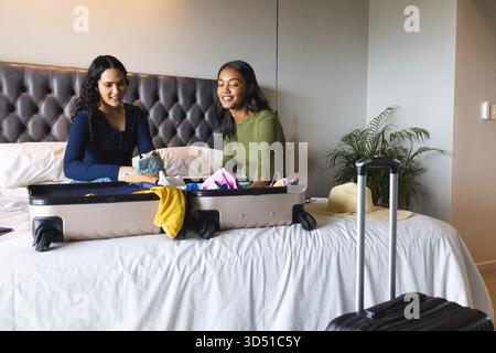 Emballage divers amis féminins pliant des vêtements colorés sur le lit à l'hôtel, avec valise et chapeau de soleil Banque D'Images