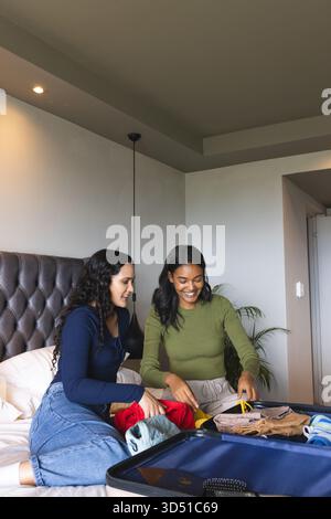 Tri de diverses amies féminines pliant et déployant des vêtements dans une valise marine sur le lit, avec la plante Banque D'Images