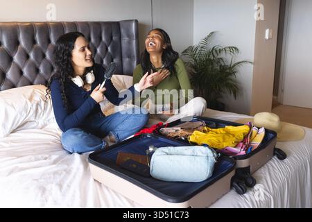 Rire diverses amies féminines tenant micro brosse à cheveux sur le lit dans la chambre moderne, avec valise ouverte Banque D'Images