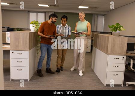 Collaboration de plusieurs collègues examinant le rapport dans l'allée de l'armoire, avec tablette, ordinateur portable et ordinateur portable Banque D'Images