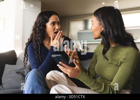 Tenant smartphone diverses amies féminines assises sur un canapé gris dans un salon moderne, avec cuisine Banque D'Images