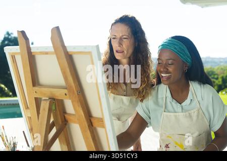 Debout diverses amies féminines étudiant la toile sur chevalet en bois dans le patio de la piscine, avec des pots de brosse Banque D'Images