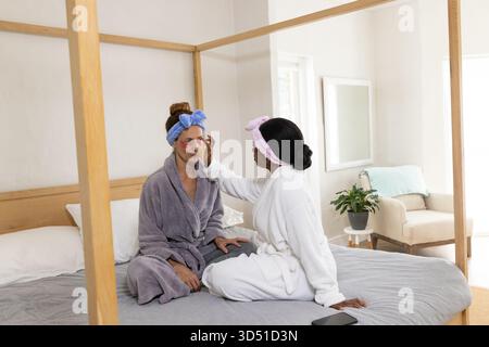 Amies féminines diverses appliquant des patchs sous les yeux dans la chambre sur un lit à baldaquin, avec téléphone Banque D'Images